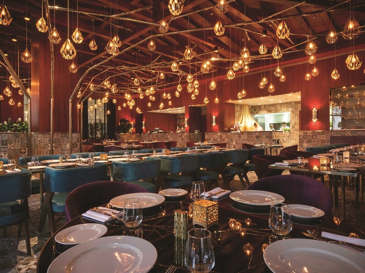 A warmly lit restaurant with velvet chairs in teal and plum, patterned tiled walls and dozens of small gold pendant lamps above the tables.