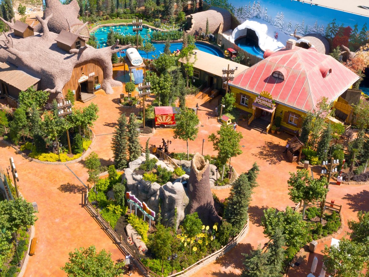 An aerial view of a themed children's land with a giant hollow tree house, a yellow cottage with a red roof, a small ride and shallow blue pools.