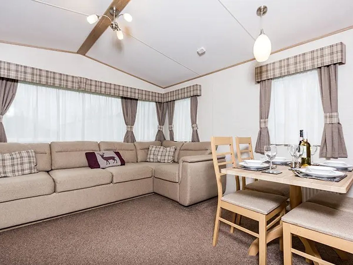 A caravan living area with a large beige corner sofa, tartan pelmets and a wooden dining table laid for four.