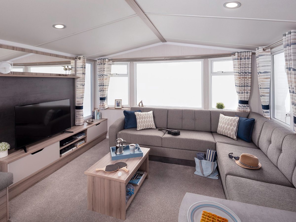 A caravan lounge with a grey wraparound sofa, blue and cream cushions, a large television on a wooden unit and a bay window.