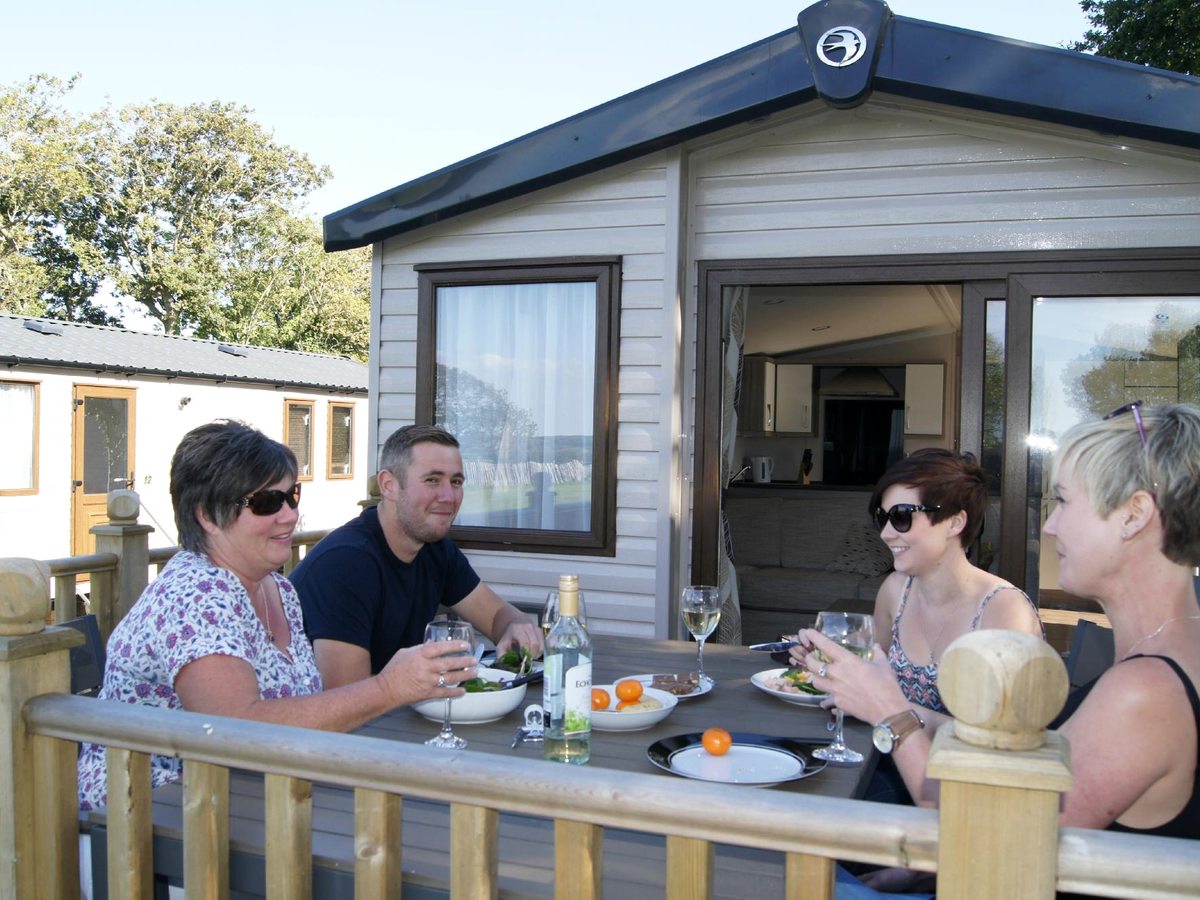 Four adults sitting at a table with plates and glasses on the timber decking outside a caravan, with its patio doors open behind.