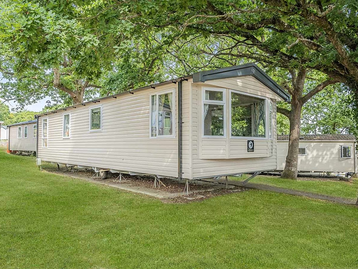 A cream static caravan with a bay window standing on a mown grass pitch under large leafy trees, with another caravan behind it.