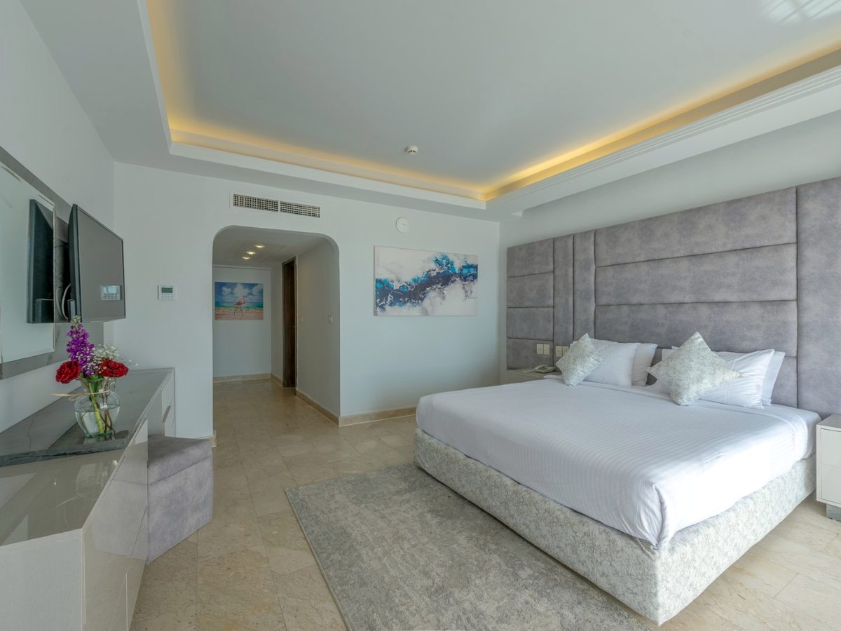 A bright bedroom with a king bed and a padded grey headboard, framed artwork, a desk and a wardrobe, with an arched hallway through to the bathroom.