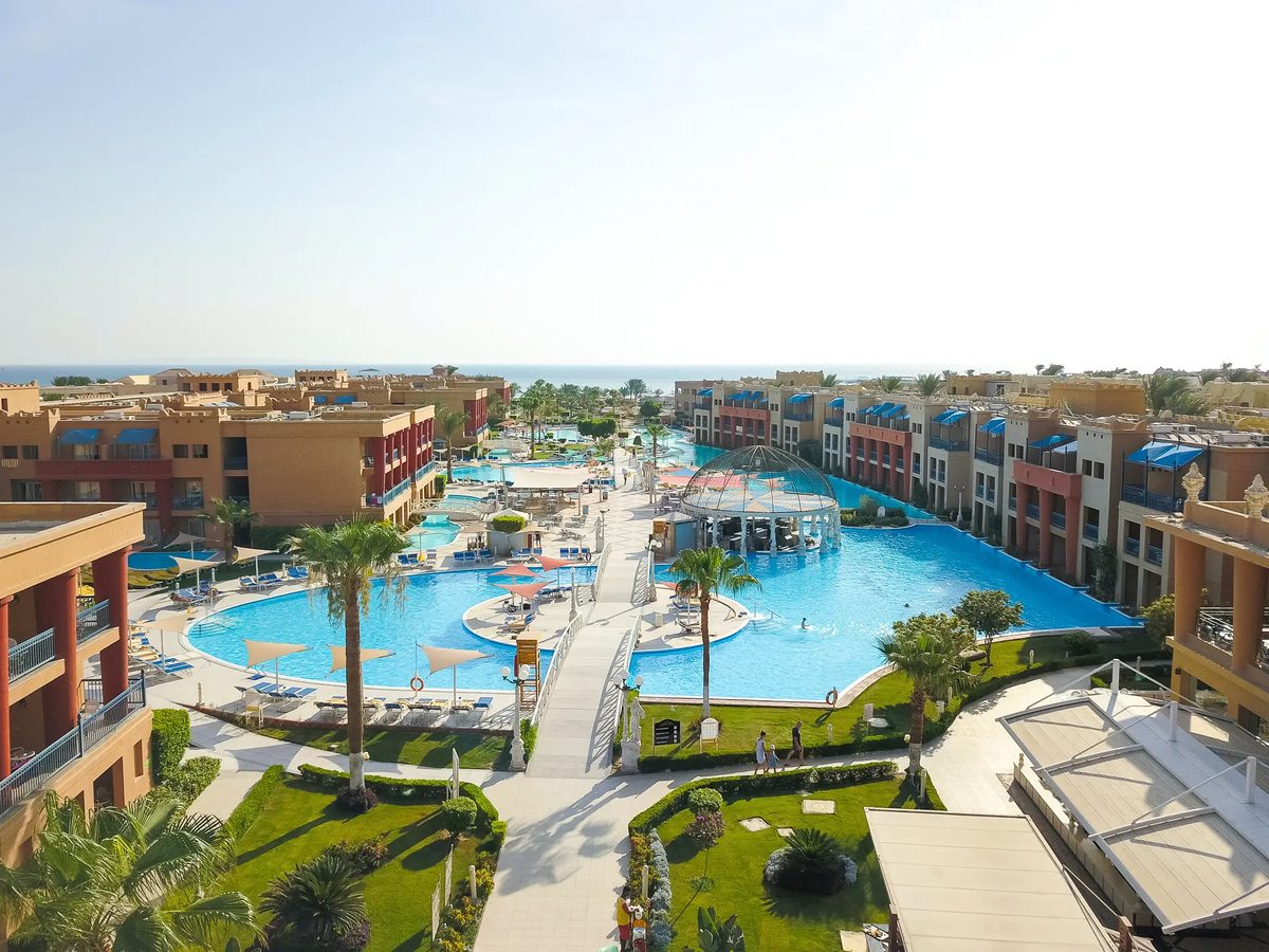 An elevated view of a large pool with a glass-domed pavilion at its centre, sun loungers and parasols around it, guest buildings on both sides and the sea beyond.
