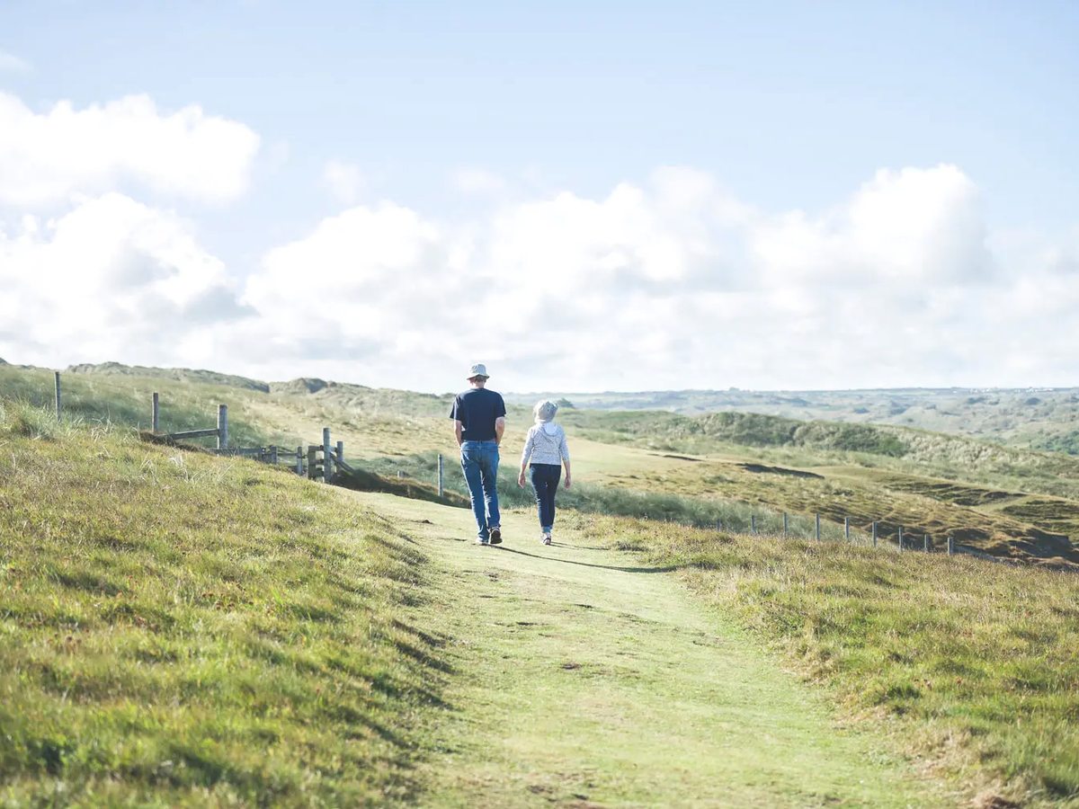 A man and a child walking a grassy path through low dunes under a bright cloudy sky.