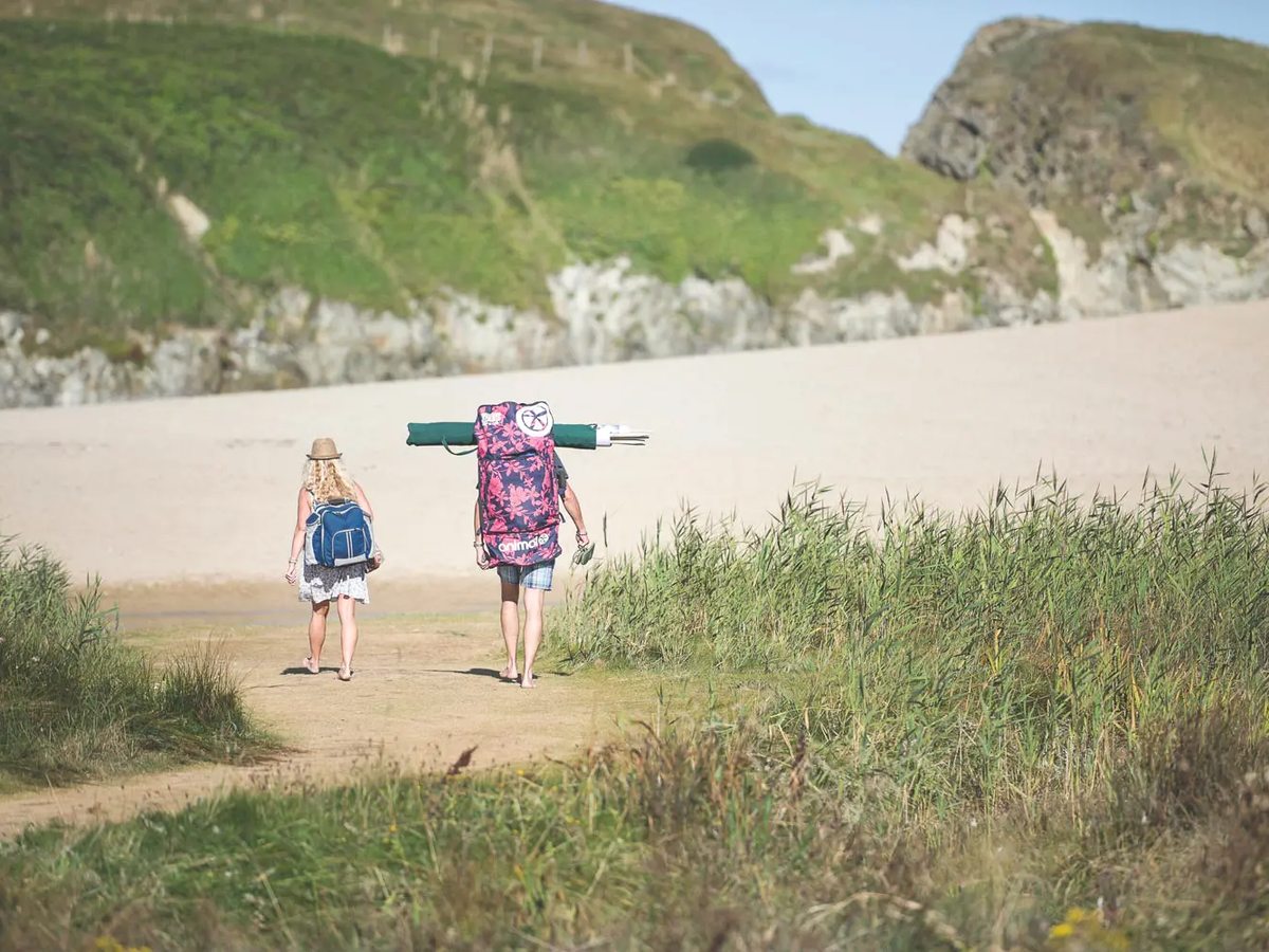 Two people carrying beach kit along a sandy path through marram grass onto a wide empty beach.