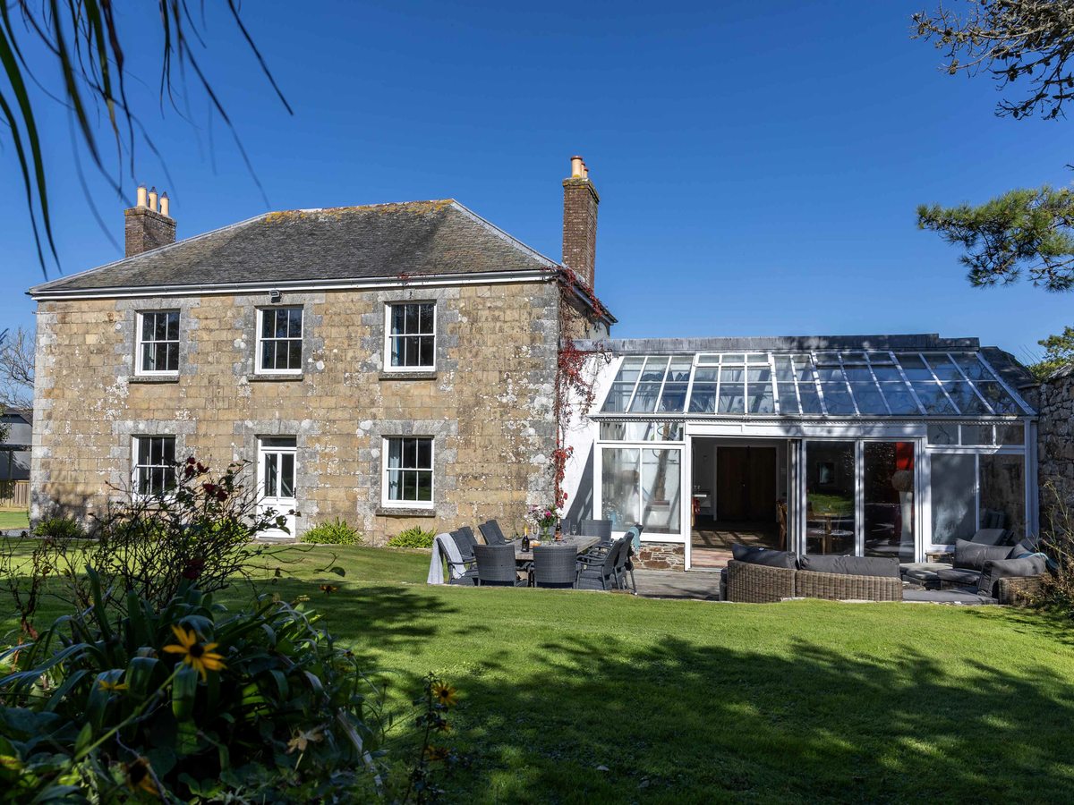 A two-storey stone farmhouse with a glass conservatory extension, outdoor dining furniture and a lawn in front.