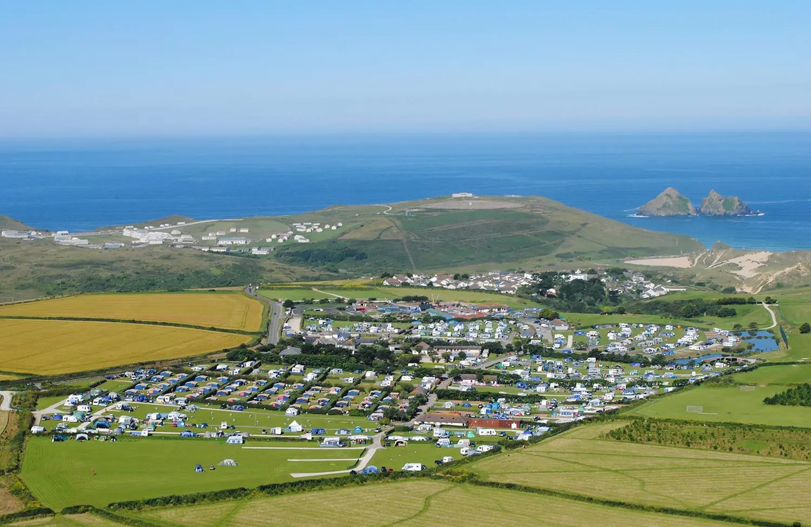 Trevornick Holiday Park from the air, its fields filled with tents and touring caravans on the slopes above Holywell Bay, with the sea and Gull Rocks beyond.