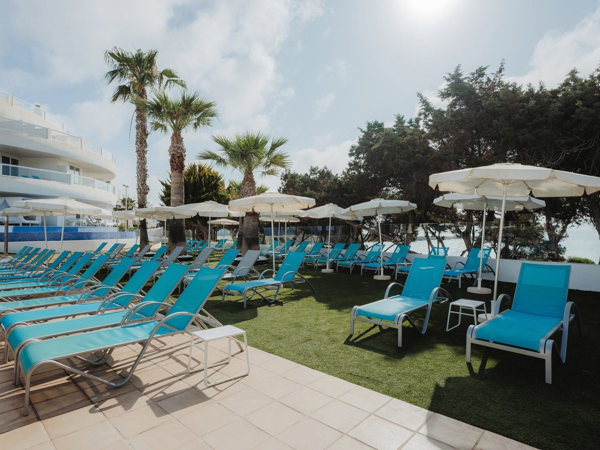 Rows of turquoise sun loungers and white parasols on a lawn and paved terrace, with palm and pine trees and the sea behind.