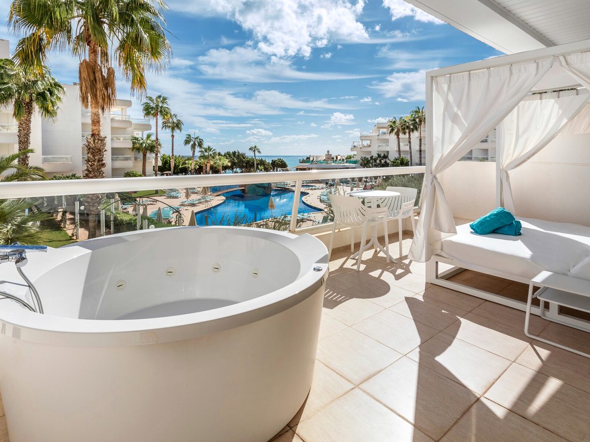 A room terrace with a round outdoor hydromassage tub, a curtained daybed and white chairs, with palms and the sea beyond.