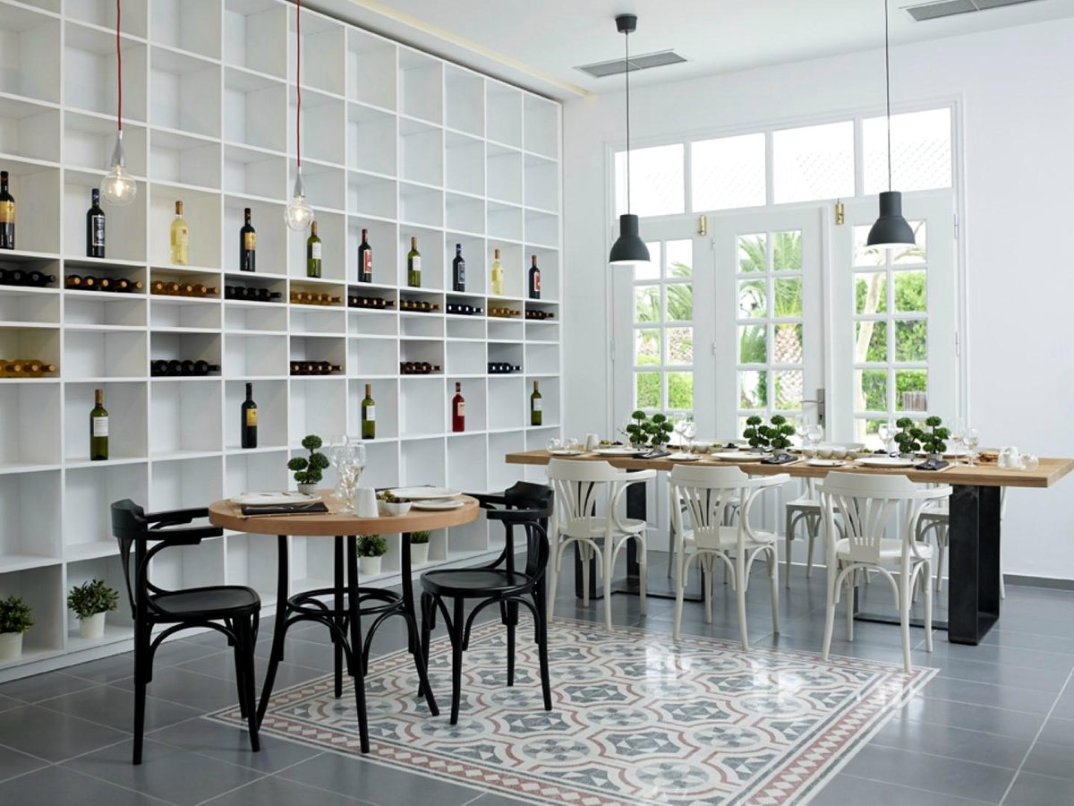 Bright white dining room with a floor-to-ceiling wine rack, round and long wooden tables, black and white bentwood chairs and patterned floor tiles.