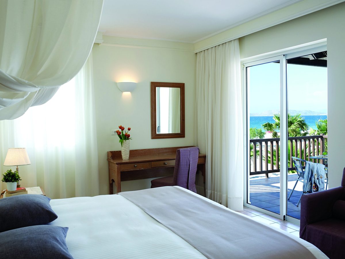 Bedroom with a double bed, wooden dressing table and mirror, and sliding doors onto a balcony with a table and chairs, palms and the sea beyond.