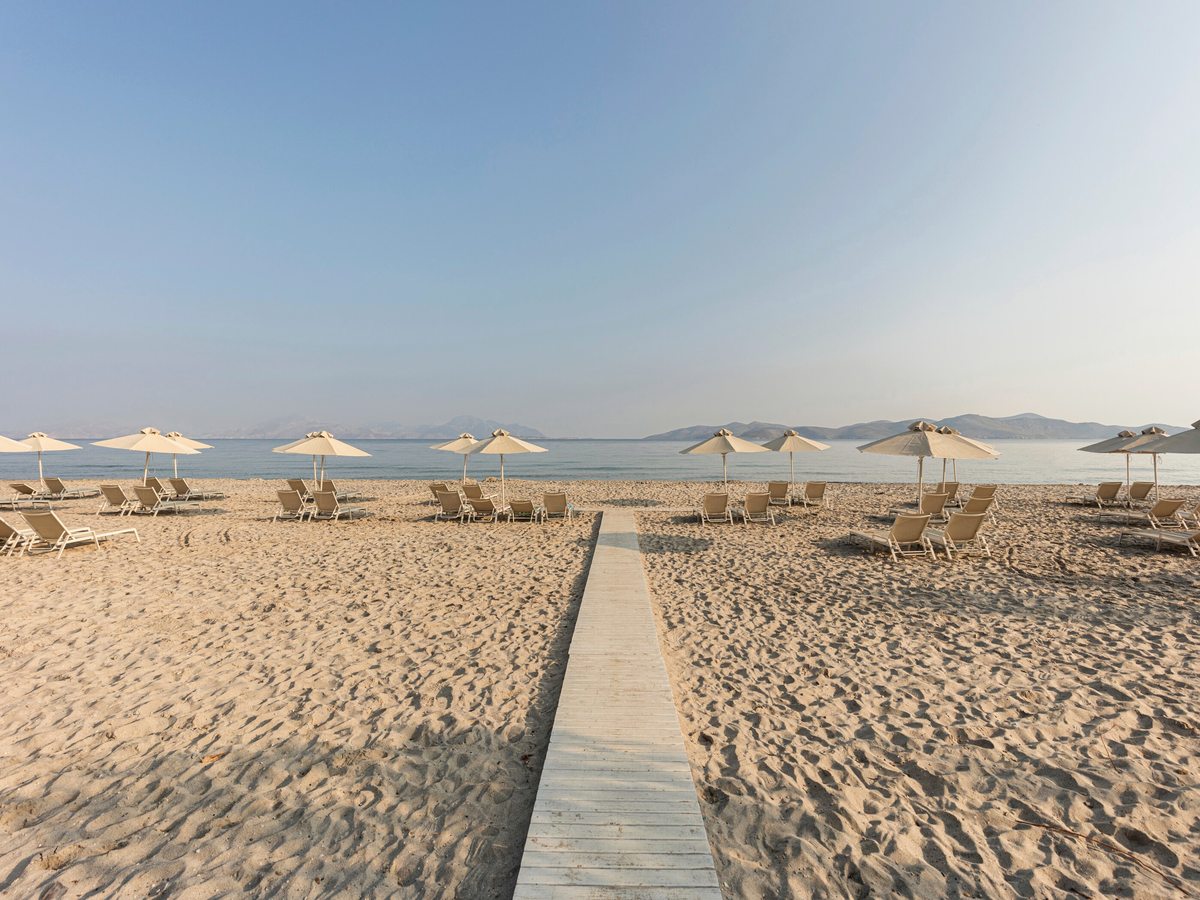 Wooden boardwalk running straight across pale sand to the water, with cream parasols and sun loungers on either side and hills across the bay.