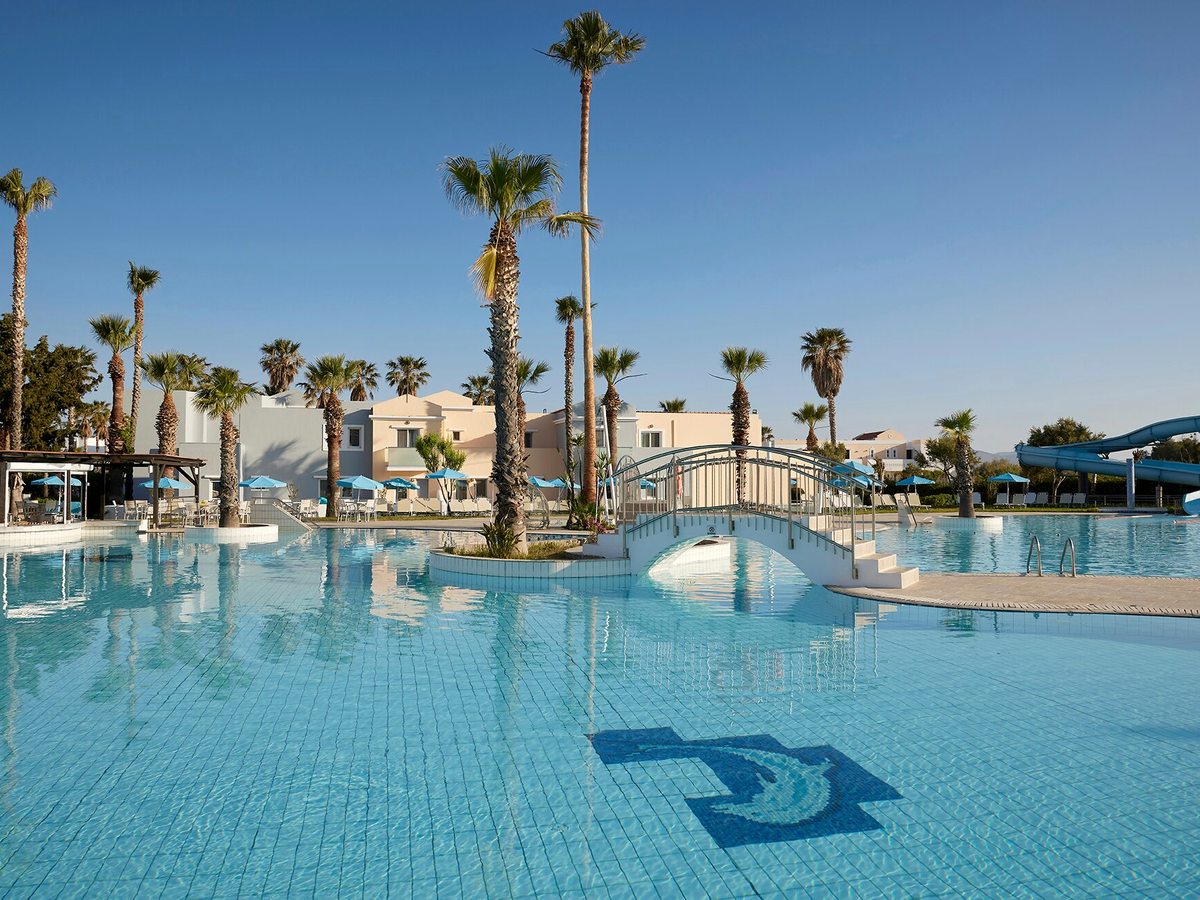 Large blue pool with a dolphin mosaic on the floor, an arched metal footbridge, palms and blue parasols, and a waterslide at the far side.