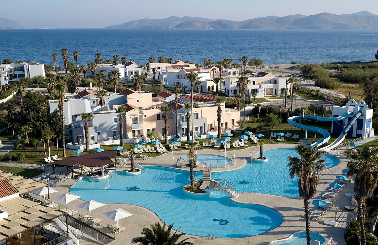 Atlantica Marmari Beach: low-rise pastel and white blocks among palms and lawns, the freeform main pool and a waterslide in front, and the sea and Turkish hills beyond.