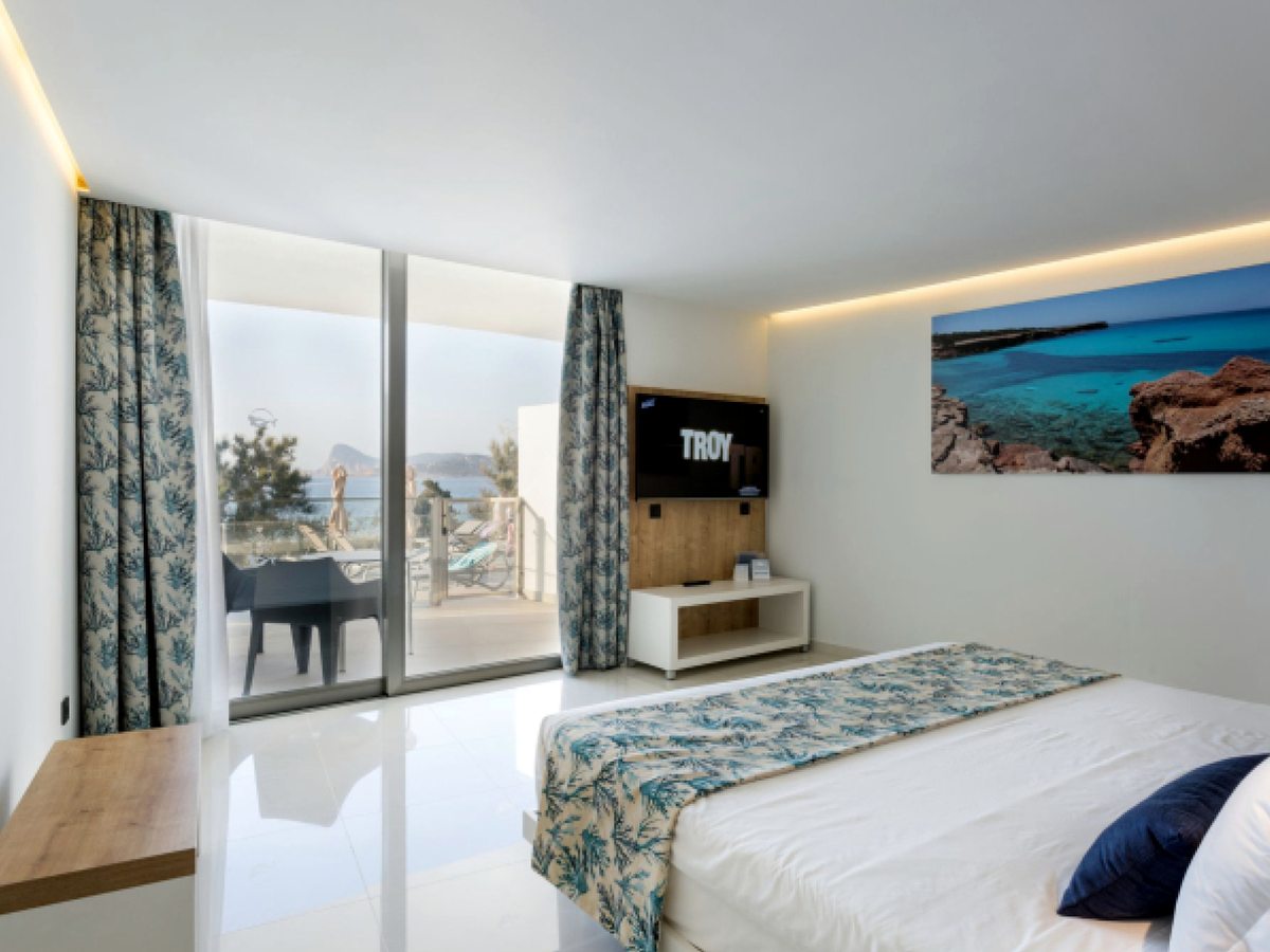 A double room with white bedding and a blue runner, a wall-mounted television and curtained windows opening to a balcony with the coast beyond.