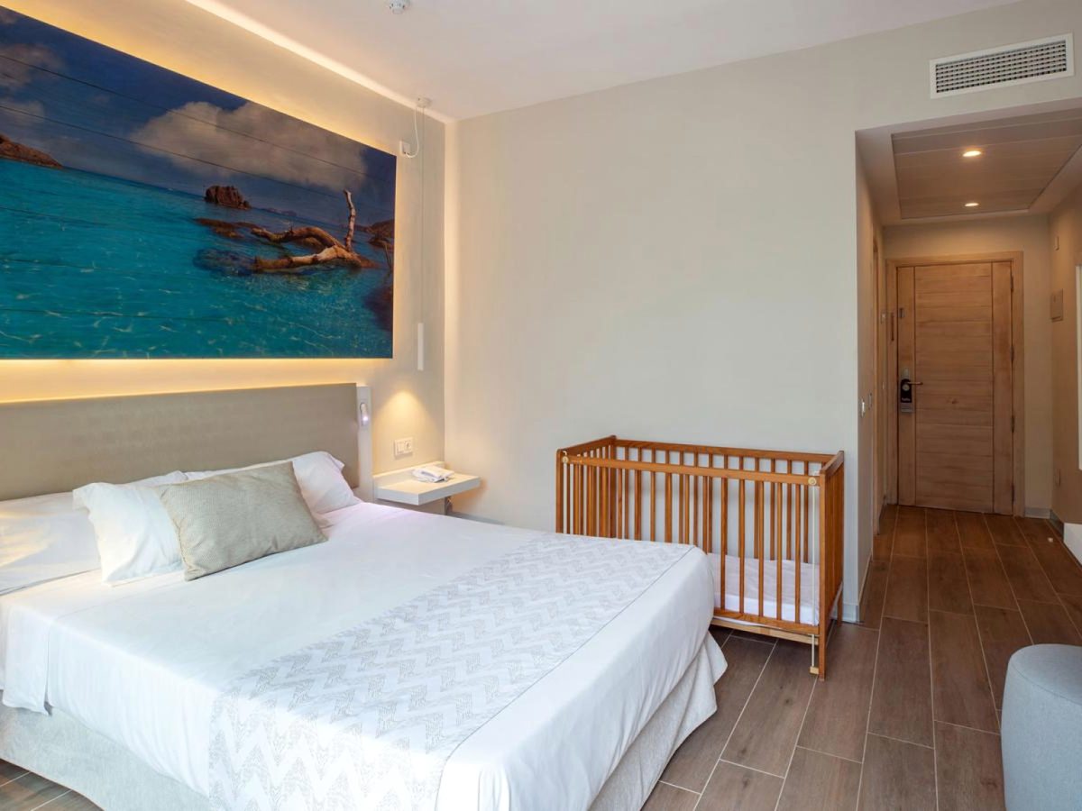 A double room with a wooden cot standing beside the bed, a lit sea photograph above the headboard and the room door beyond.