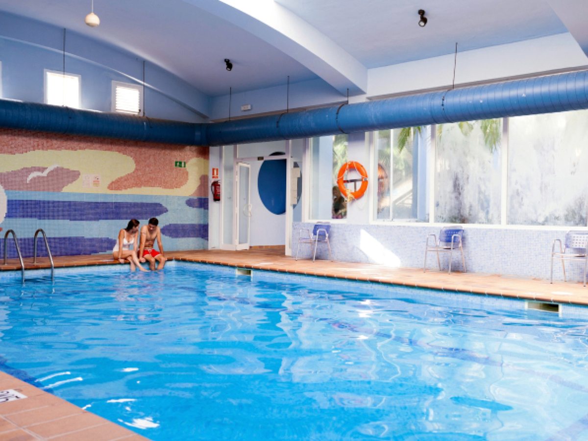 An indoor pool in a room with a painted seaside mural, poolside chairs and windows down one side.