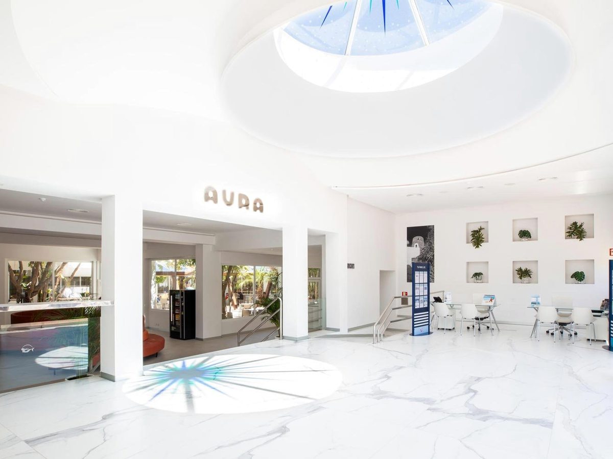 A bright white lobby with a circular skylight, a pale marble floor, AURA lettering on the wall, glass entrance doors and lounge seating.