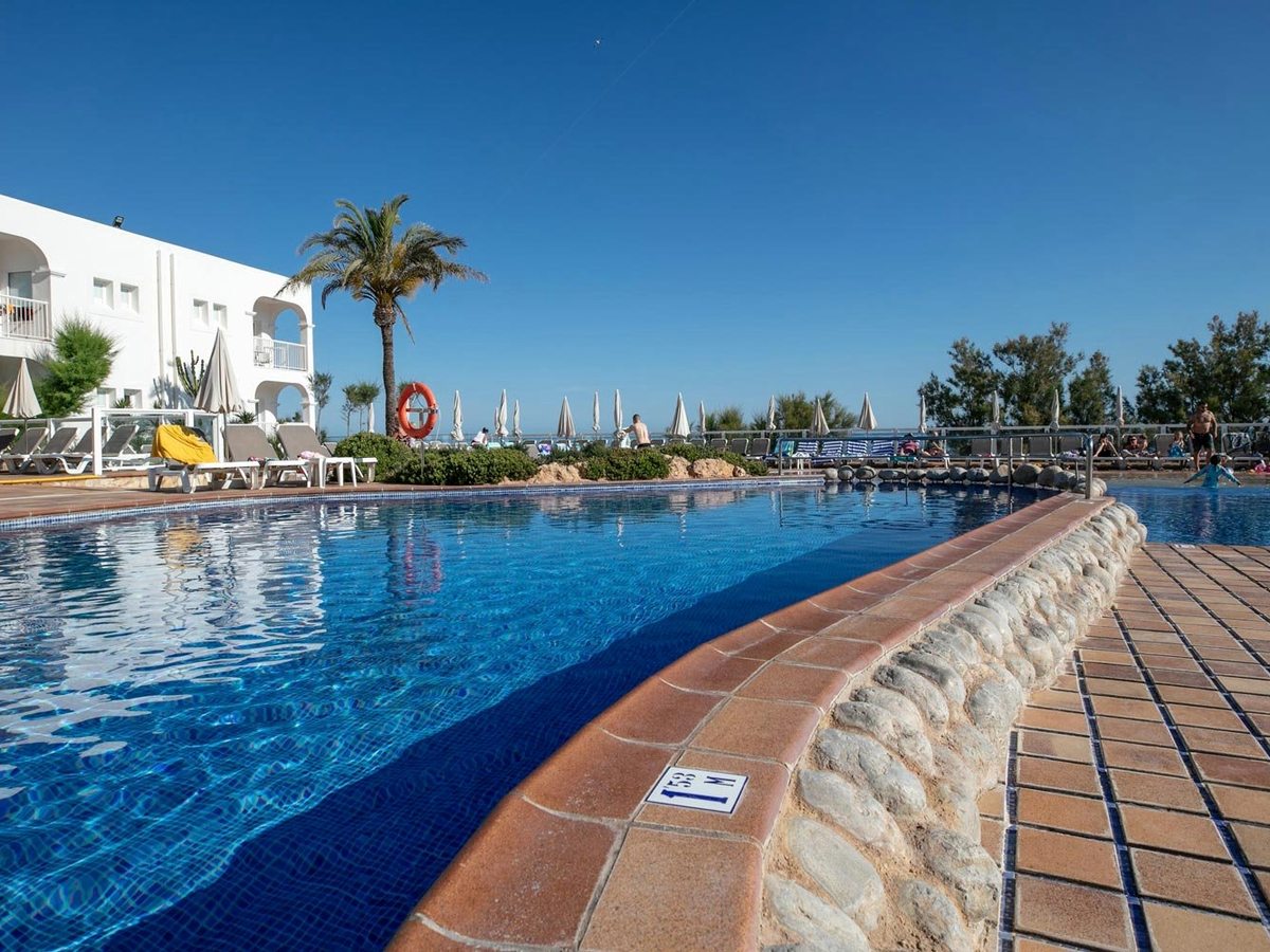 A large outdoor pool curving past a stone-edged deck and sun loungers, a depth marker on the tiled edge and the white resort building and palms behind.