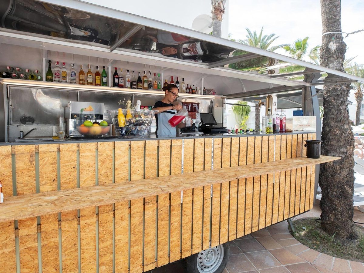 A wheeled outdoor drinks trailer under palm trees, clad in timber boards, its counter set with spirit bottles, cut fruit and a till, and a barwoman serving.
