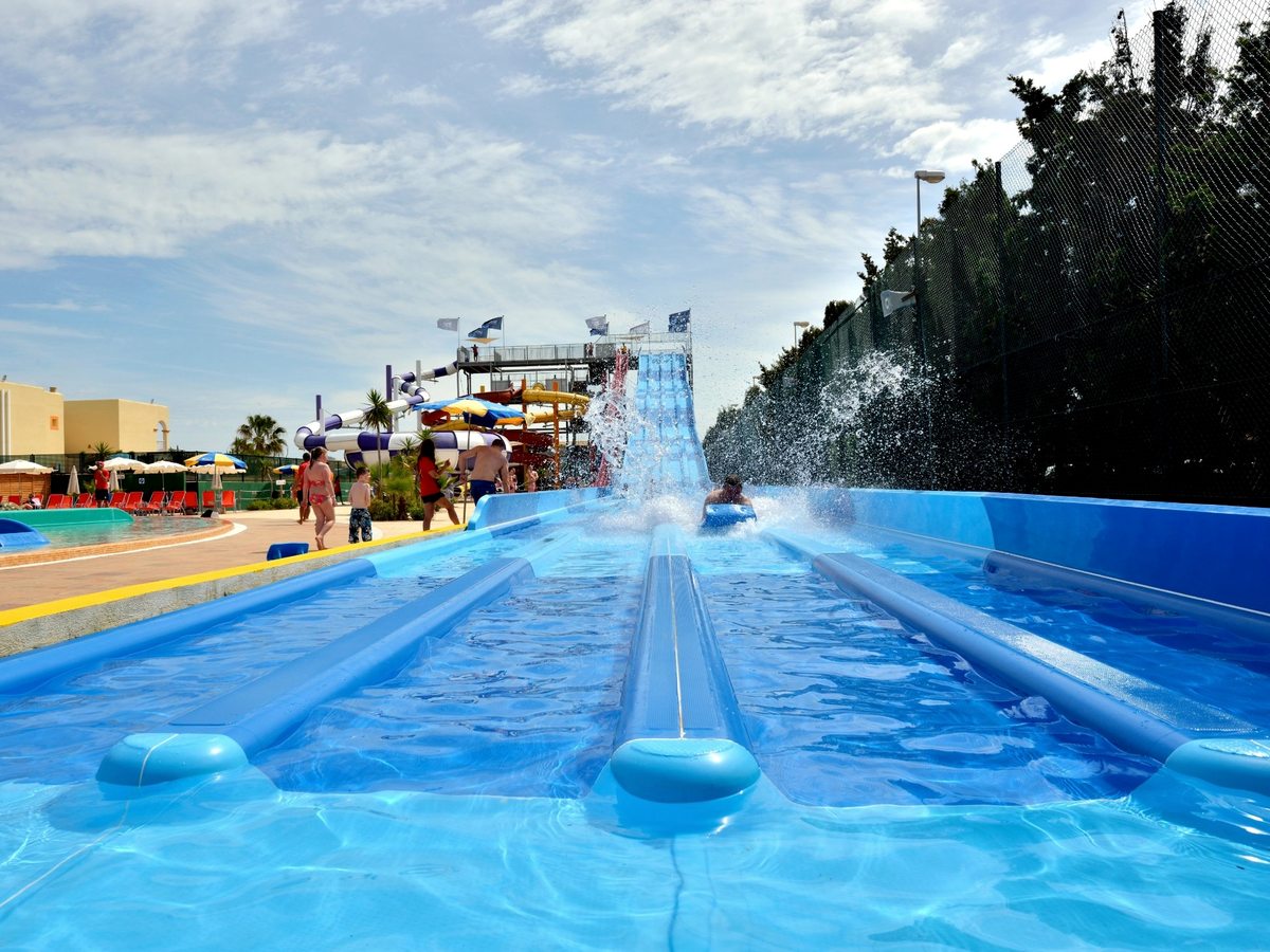 A wide multi-lane mat racing slide with a rider splashing down at the bottom, and the tube-slide tower and pools alongside.