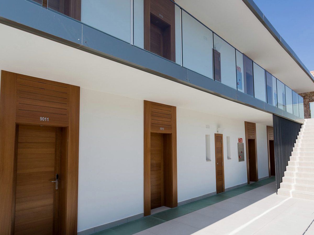 The outside of an accommodation block with numbered wooden room doors along a walkway, glass-fronted balconies above and a flight of stone stairs to the upper floor.