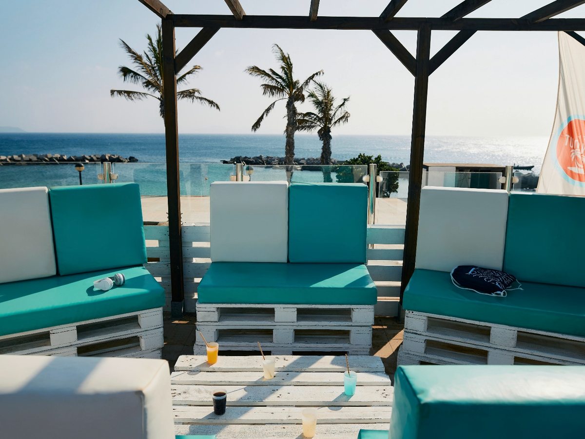 Turquoise and white cushioned pallet sofas under a timber pergola on a terrace, with palm trees, a rock breakwater and the sea behind.