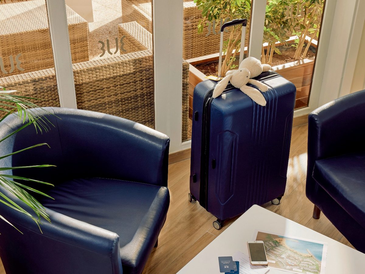 A reception seating area with navy tub armchairs around a low table of travel brochures, a suitcase and a child's soft toy beside a window.
