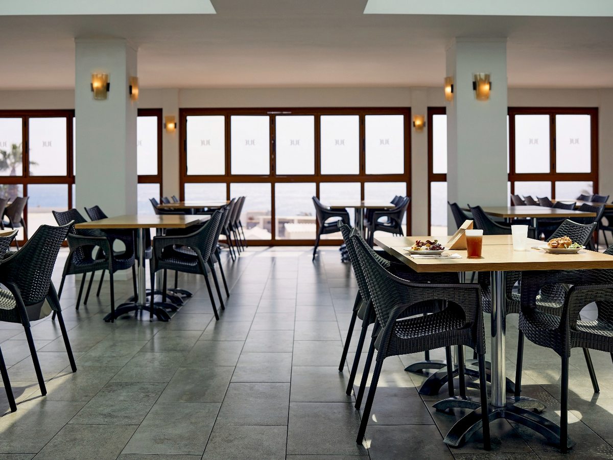 An indoor restaurant dining room with square tables, dark woven chairs, a tiled floor and tall windows along one wall.