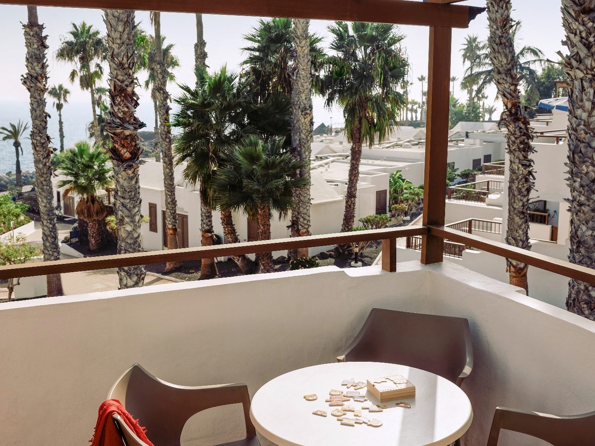 A balcony with a round white table and moulded chairs, framed by palm trees, with the resort's white bungalow roofs and the sea beyond.