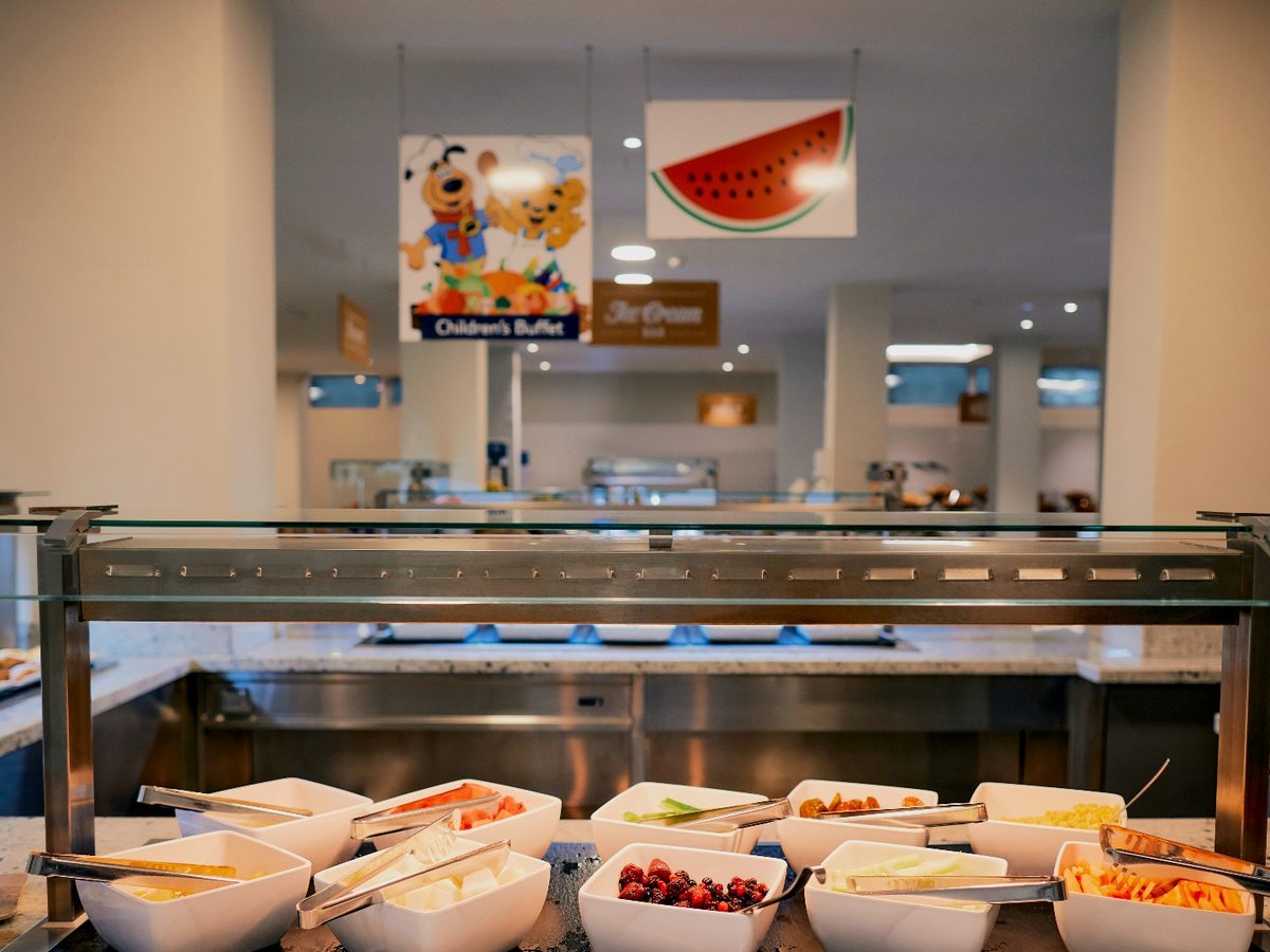 A self-service buffet counter under a glass guard, with white bowls of fruit and salad below a cartoon children's buffet sign.