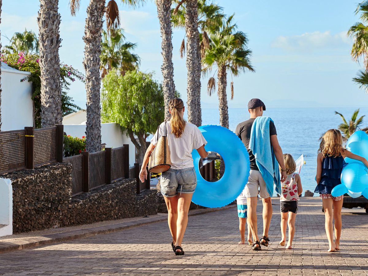 A family carrying inflatable rings and towels walking down a paved, palm-lined path towards the sea.