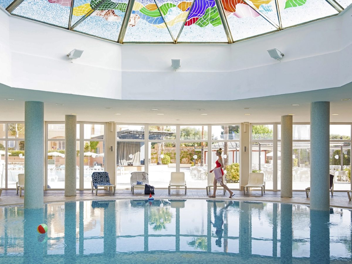 An indoor pool below a stained-glass skylight, with white columns, loungers along the side and glass doors to the terrace outside.