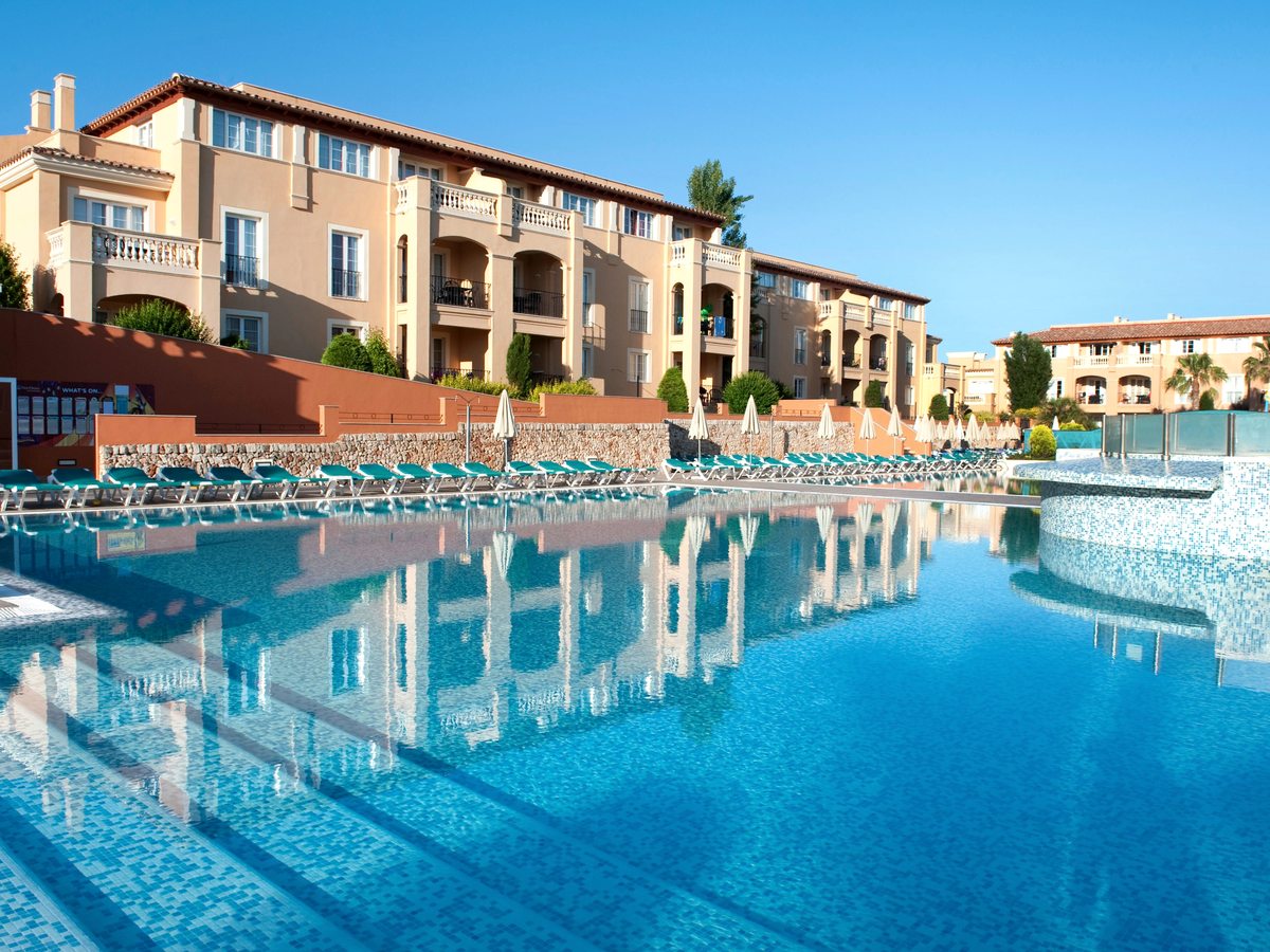 Three-storey cream apartment blocks with arched balconies behind a stone retaining wall, with rows of turquoise loungers and folded parasols along the main pool below.