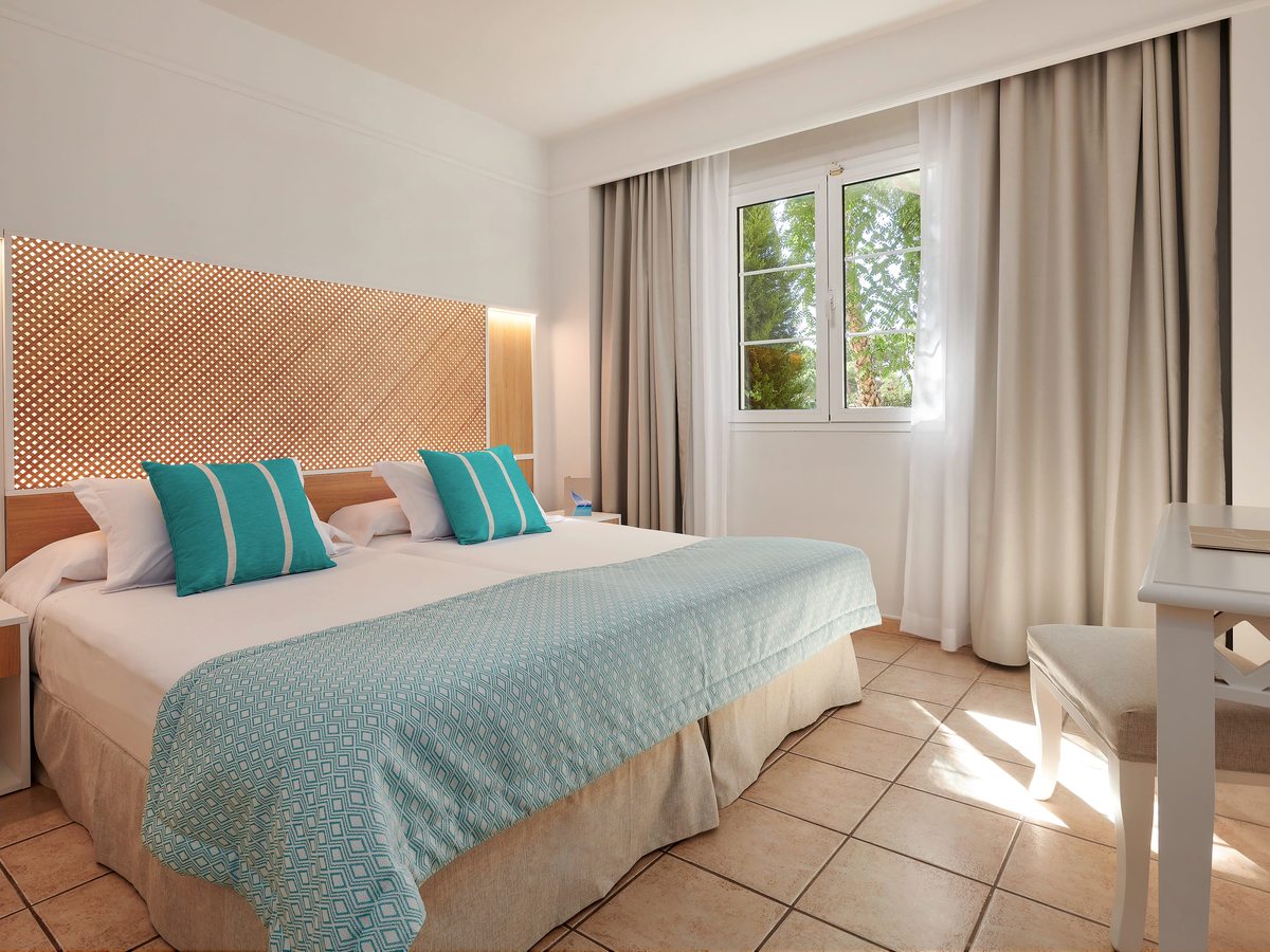 A bedroom with two single beds pushed together under a patterned wooden headboard, turquoise cushions and a turquoise throw, a window onto greenery and a small desk and stool.