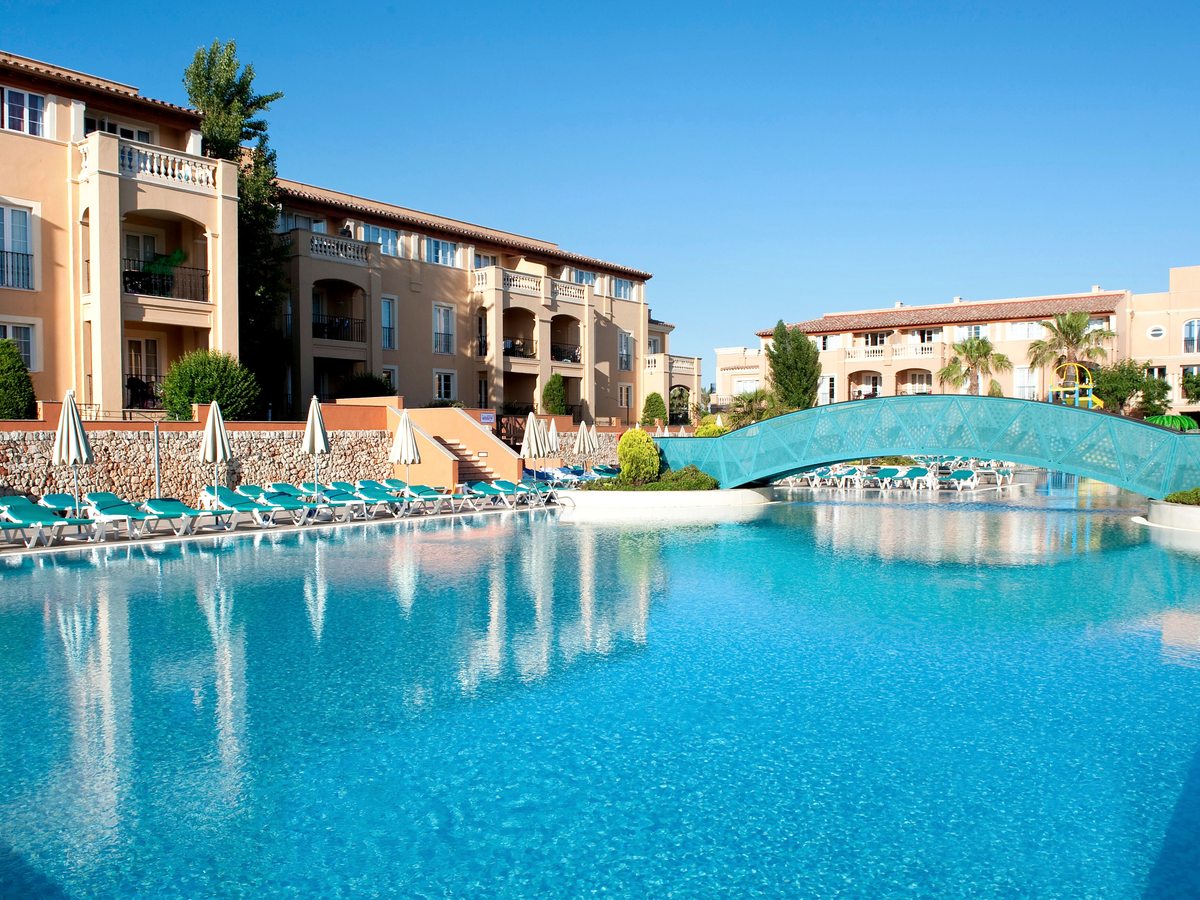 A large curved outdoor pool with a turquoise arched footbridge crossing it, rows of loungers and parasols along the edge and cream apartment blocks behind.