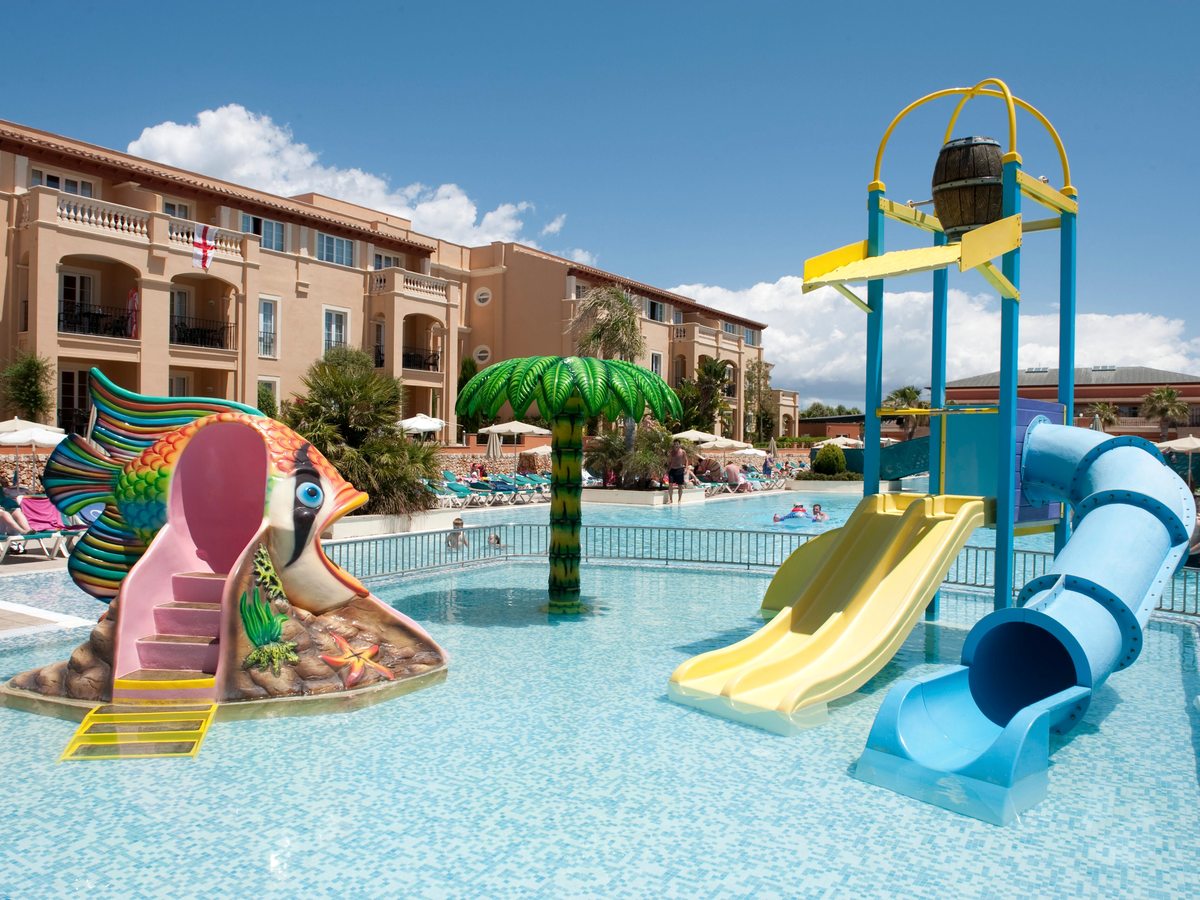 A shallow children's pool with a yellow open slide and a blue tube slide from a play tower with a tipping bucket, a fish-shaped play structure and a palm-tree water feature.