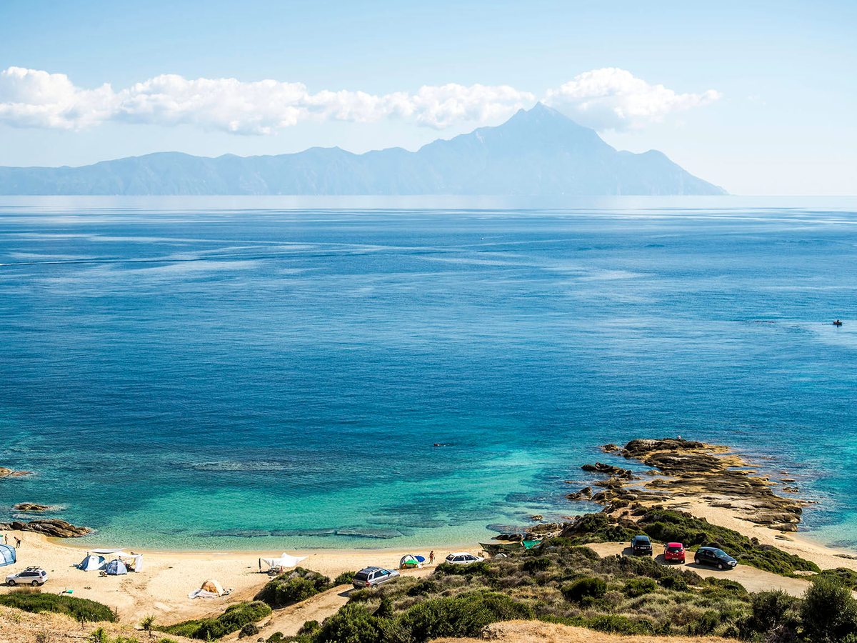 A sandy beach at the foot of green hills with clear turquoise water, and a mountain rising across the water in the distance.