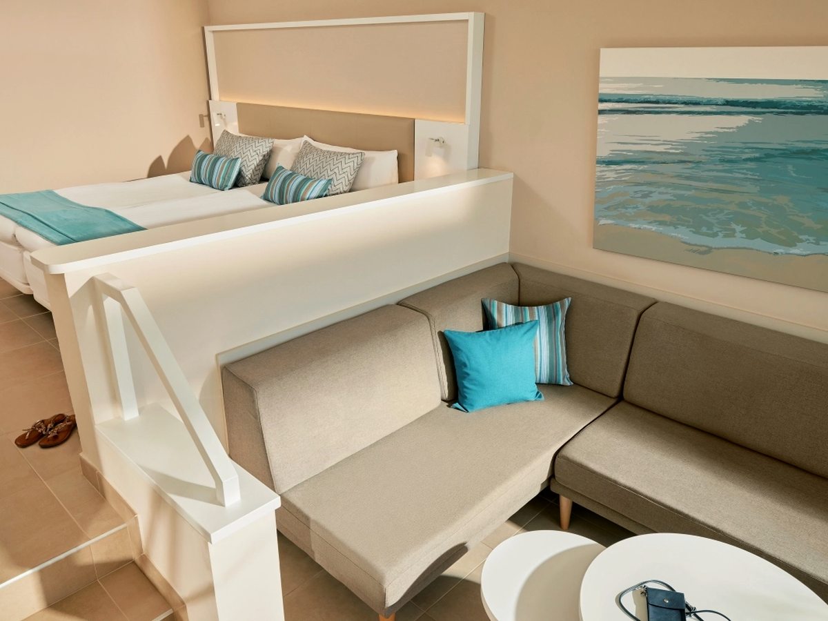 Twin beds on a raised platform behind a white rail and short flight of steps, with an L-shaped sofa bed and low round tables on the level below and a painting of breaking waves on the wall.