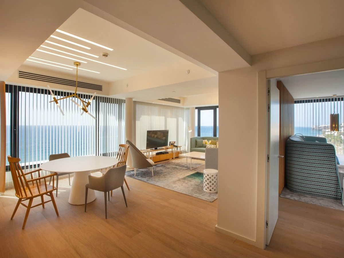 An open-plan suite with a round white dining table and wooden chairs by full-height sea windows, a sofa and television beyond and a bedroom through an open door.