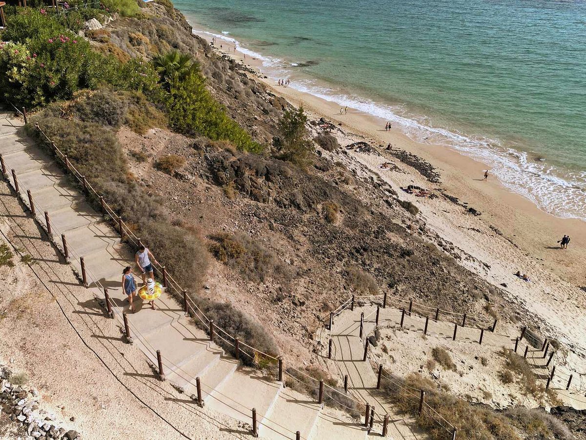 Concrete steps and a rope-and-post handrail zigzagging down a scrubby cliff to a sandy beach, with two adults and a child carrying a yellow ring on the way down.