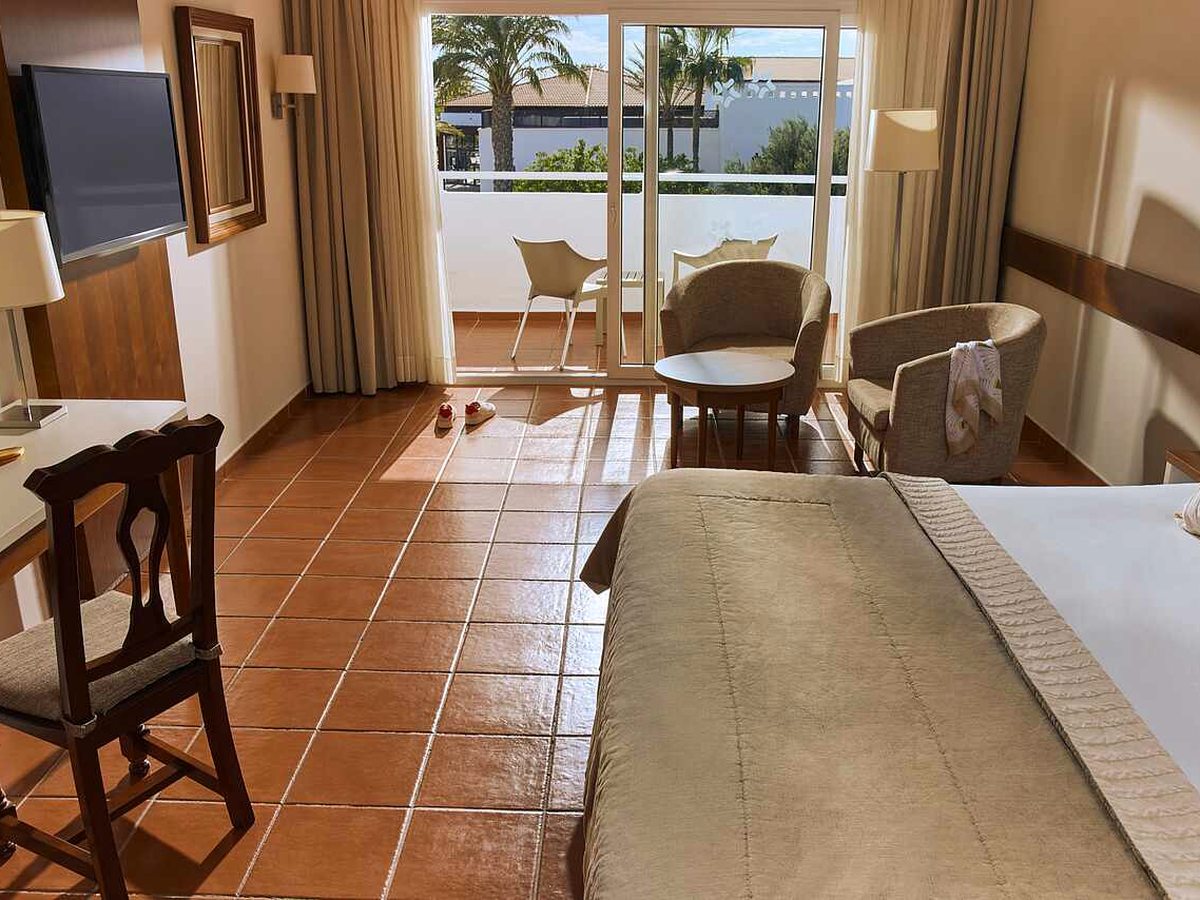 Bedroom with a double bed, armchairs and a coffee table by open balcony doors, with palms and the resort gardens outside.