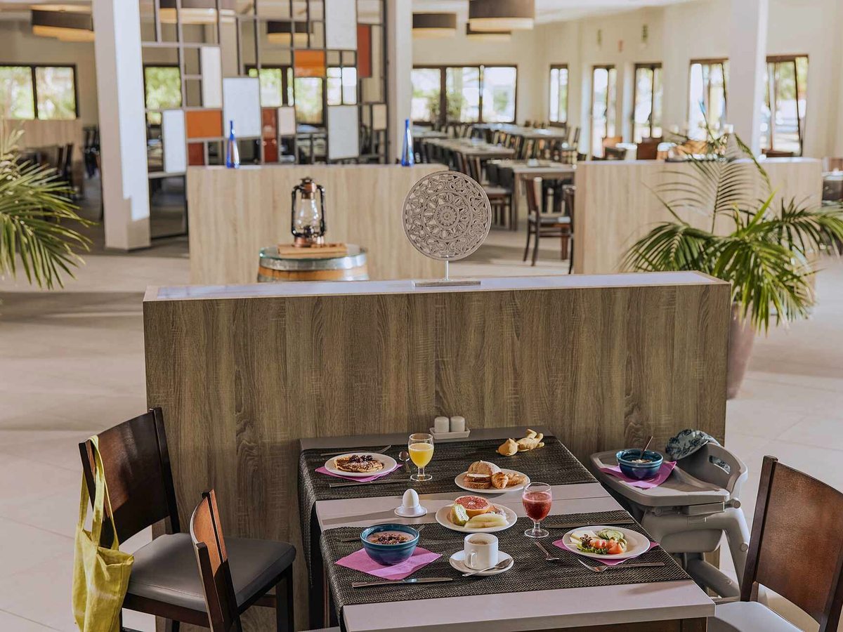Buffet restaurant with a laid breakfast table of pancakes, fruit and coffee in the foreground and a high chair beside it, with pale timber serving counters and further tables behind.