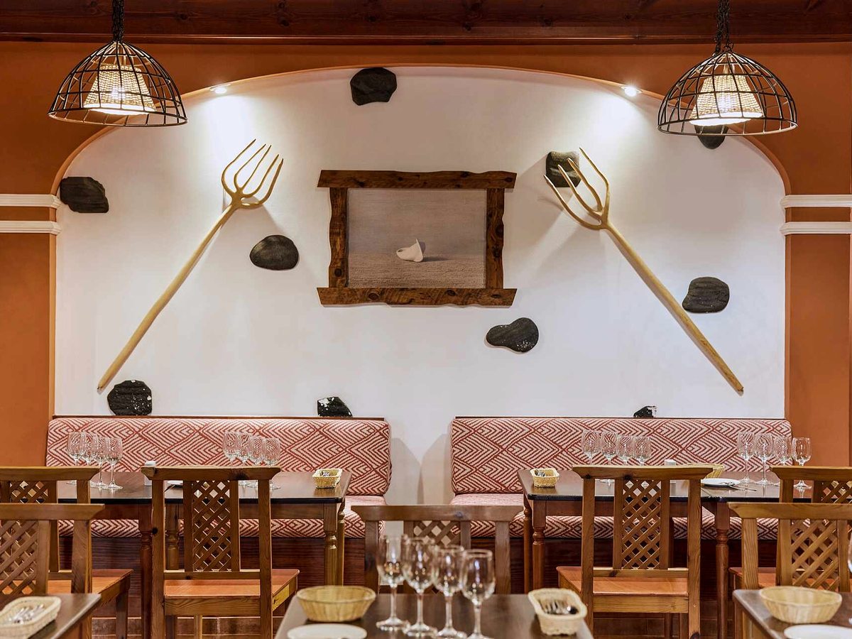 Rustic dining room with a whitewashed arch set with volcanic stones and wooden farm tools, red and white banquette seating, wooden chairs and tables laid with wine glasses.