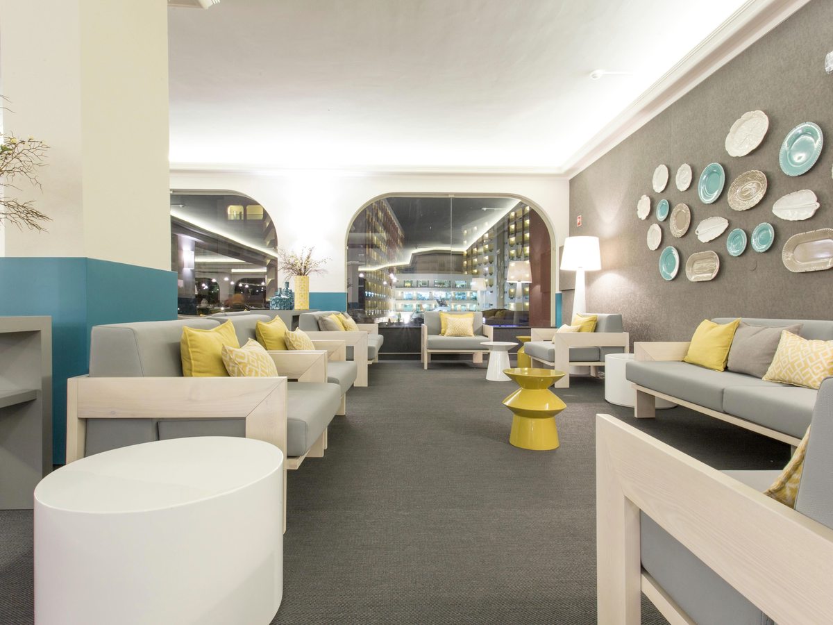 Lobby lounge with pale grey sofas, yellow cushions, round side tables and a wall hung with decorative plates, the lit bar behind.