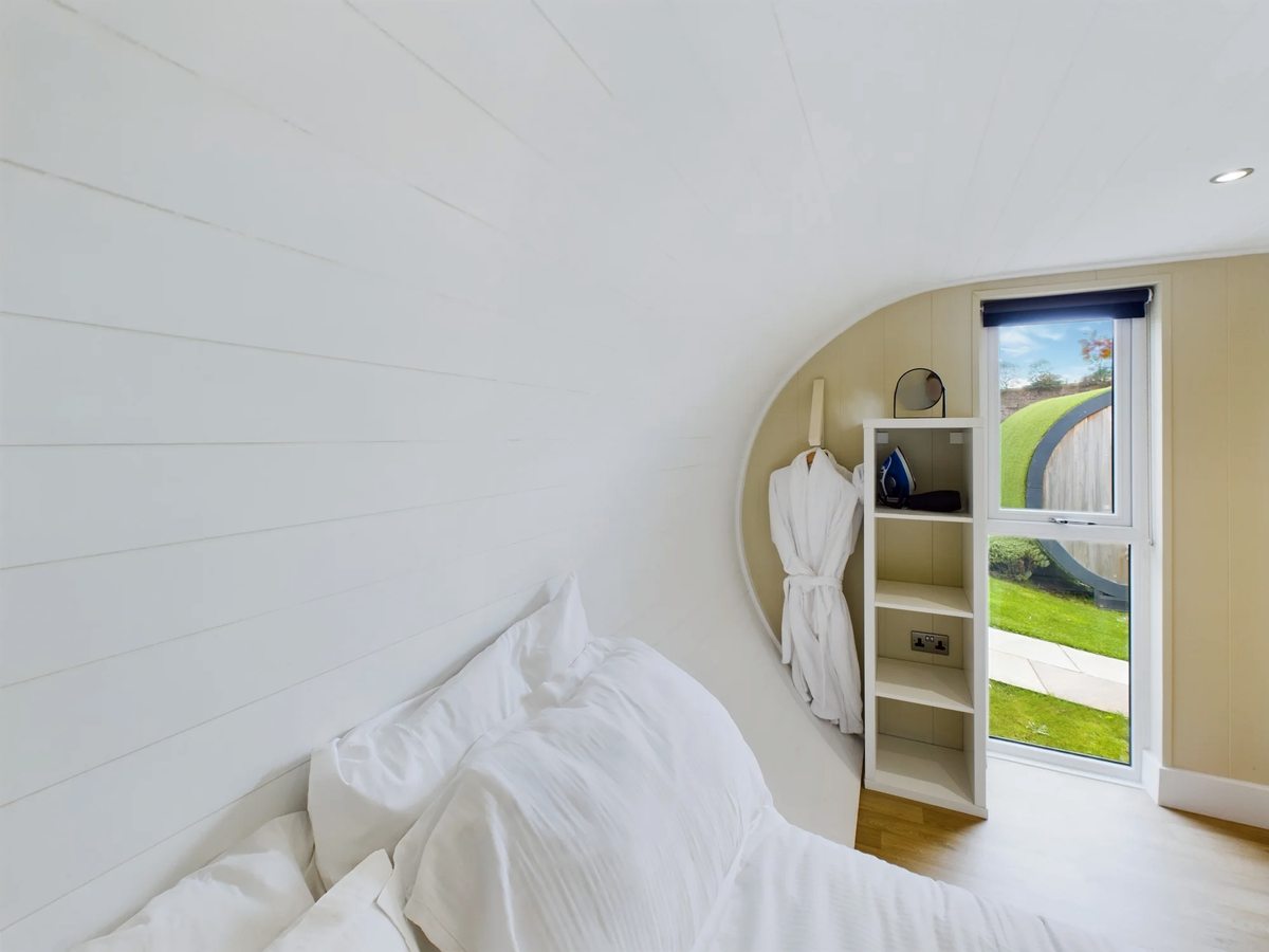 The double bedroom in a lakeside pod, with white bedding under the pod's curved white ceiling, open shelving, a hanging robe and a full-height window onto the grass outside.