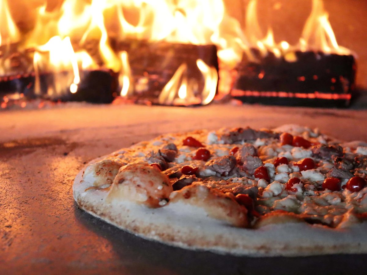 A stone-baked pizza topped with meat cooking on the hearth of a wood-fired oven, with logs burning behind it.