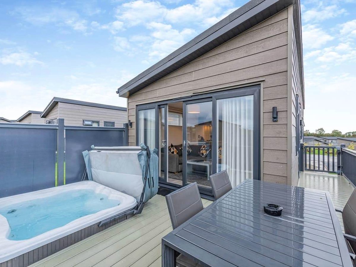 A bubbling hot tub on a lodge's timber deck beside an outdoor dining table, with the lodge's glazed doors behind.