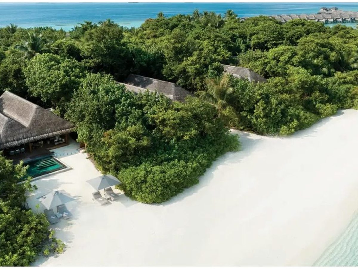 Aerial view of dense green tree canopy covering low thatched-roof villas, with a broad sweep of white sand, sun loungers and parasols at the water's edge.